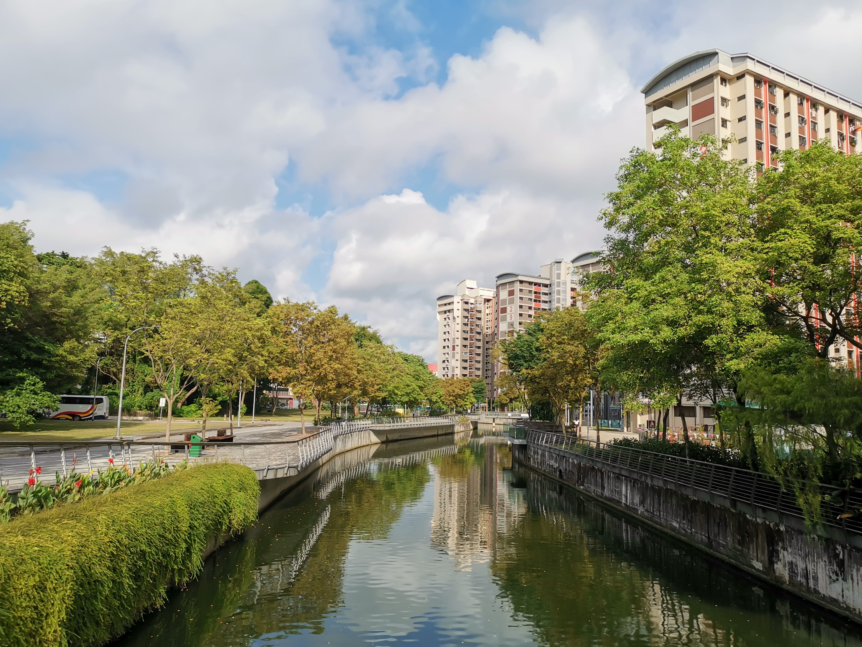 A narrow river lined with greenery and trees, with tall apartment buildings in the background under a partly cloudy sky.